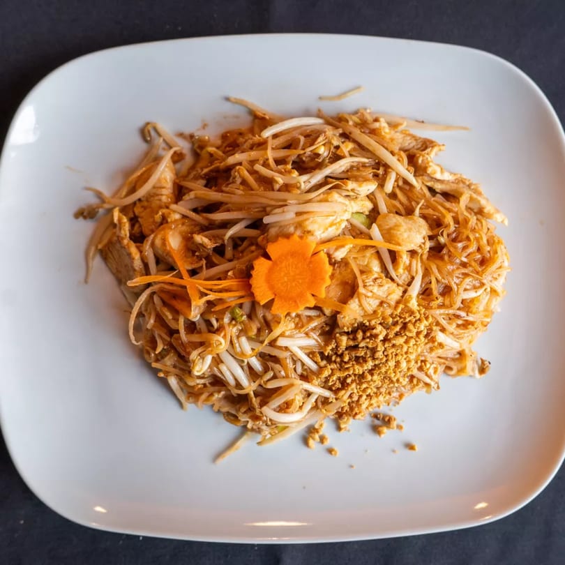 Pad Thai noodles with chicken, bean sprouts, crushed peanuts, and a carrot garnish on a white plate.