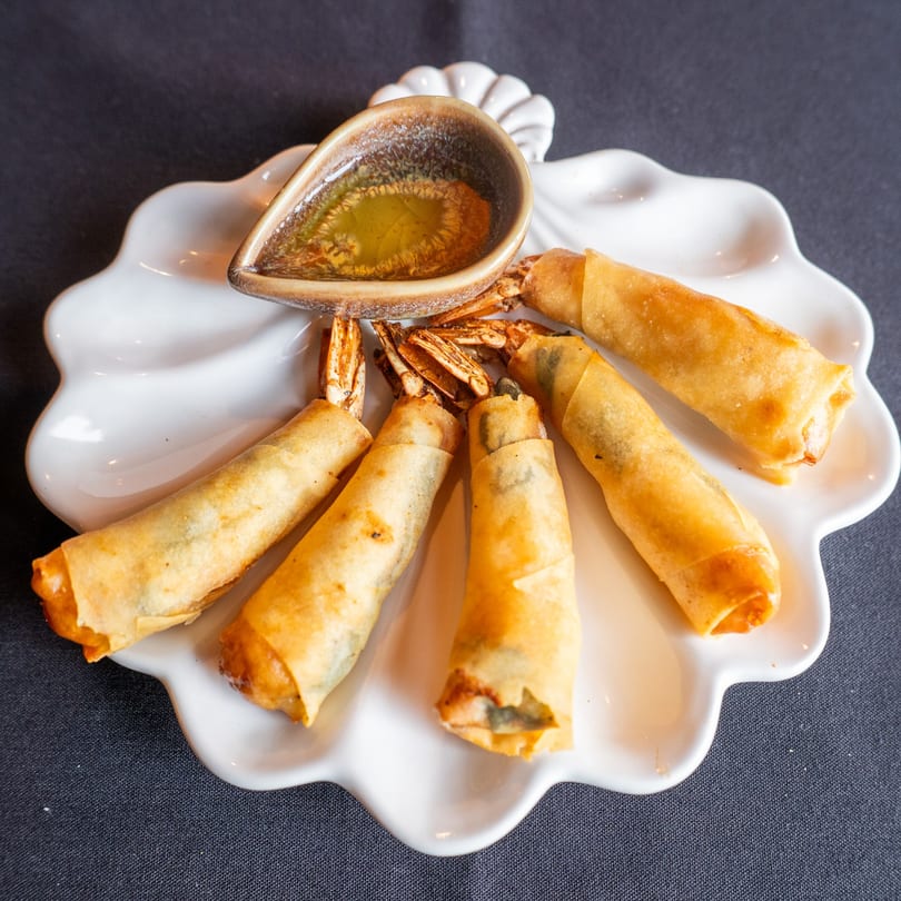 Six golden-fried shrimp spring rolls with tails visible, arranged around a small dish of dipping sauce.