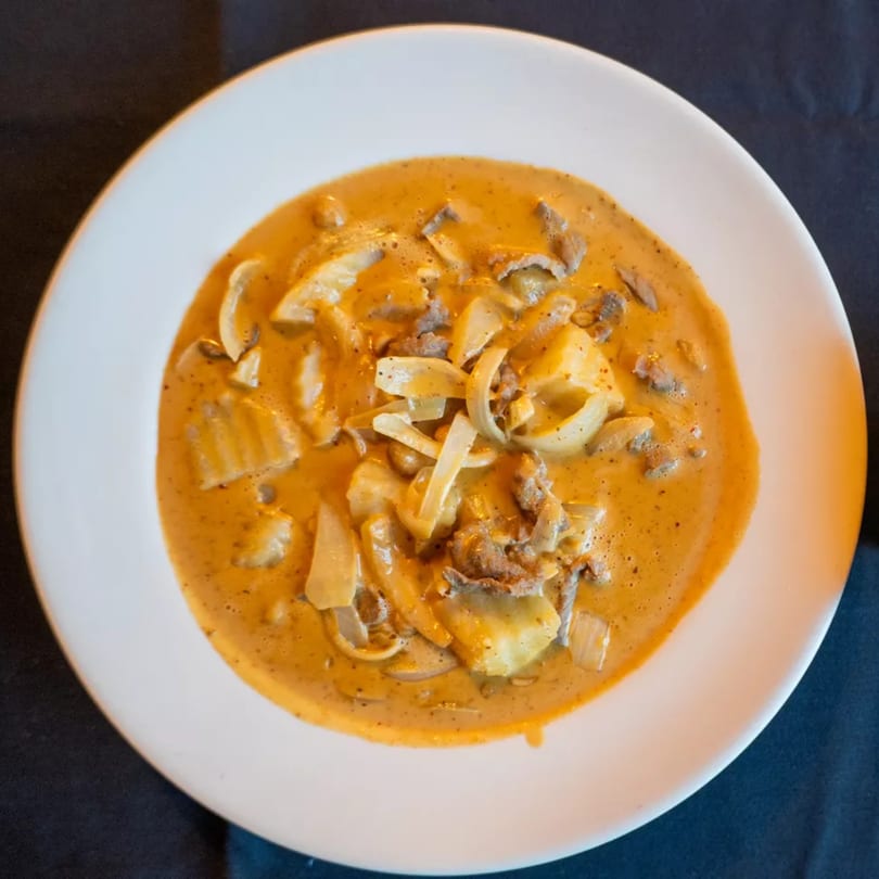 Creamy orange curry with sliced onions, potatoes, and beef in a white bowl on a dark surface.