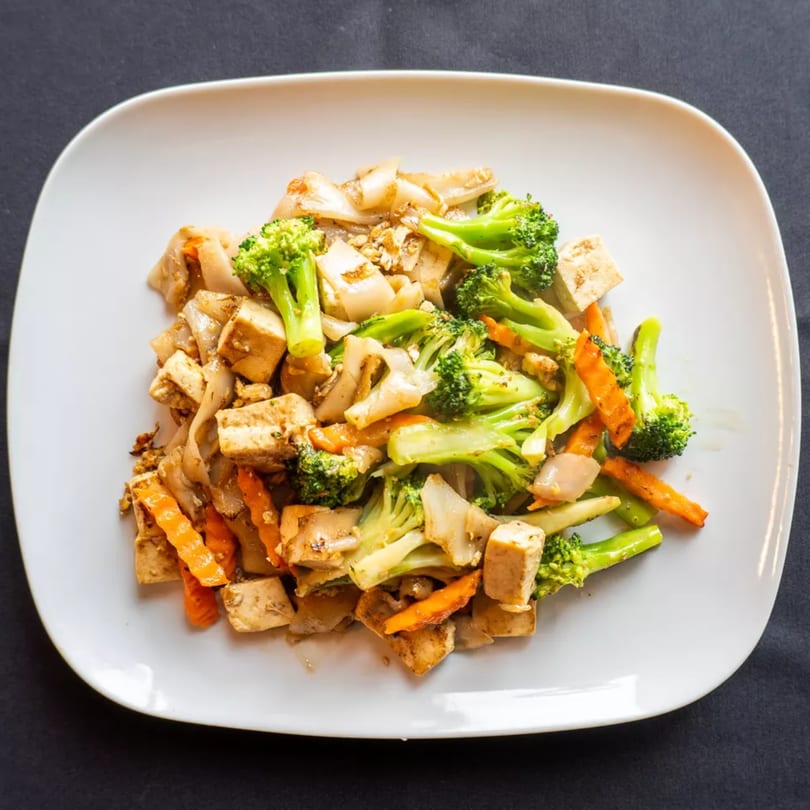 A white plate holds a serving of Pad See Ew with tofu, broccoli, and carrots.